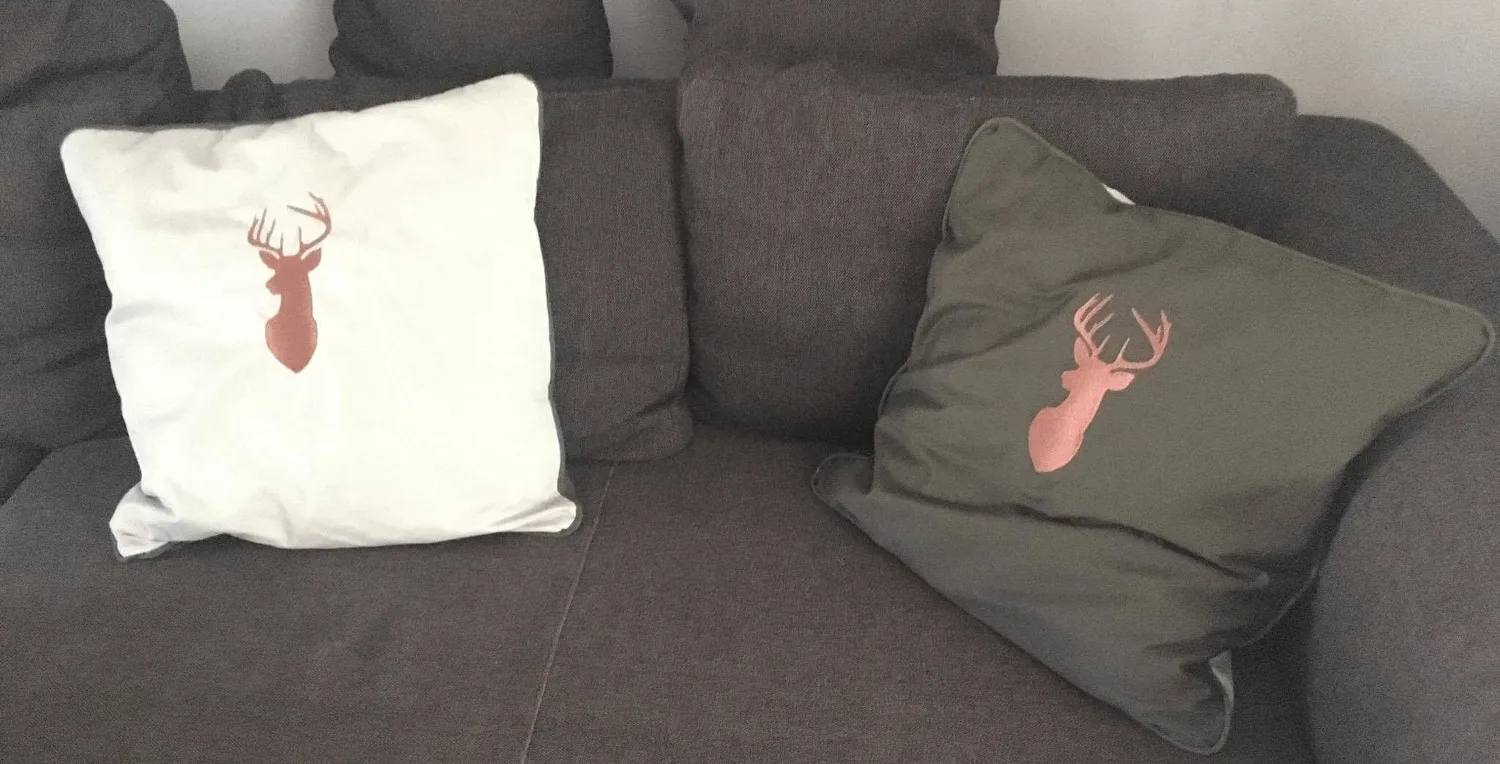 Zwei Kissen mit gesticktem Hirschmotiv in Cremeweiß und Olivgrün auf einem grauen Sofa.