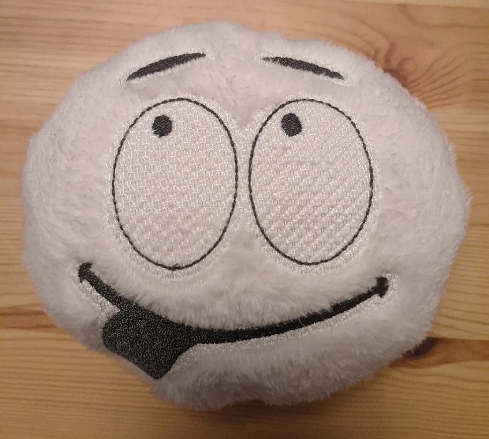 Rundes Stoffspielzeug aus weißem Plüsch mit gesticktem Gesicht auf Holztisch.