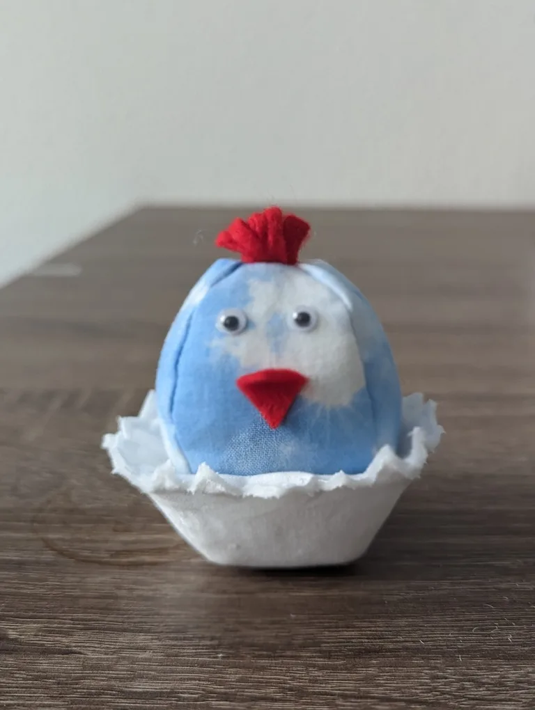 Kükenfigur aus blauer Baumwolle in weißer Eierschale mit roten Filzdetails und Wackelaugen auf Holz