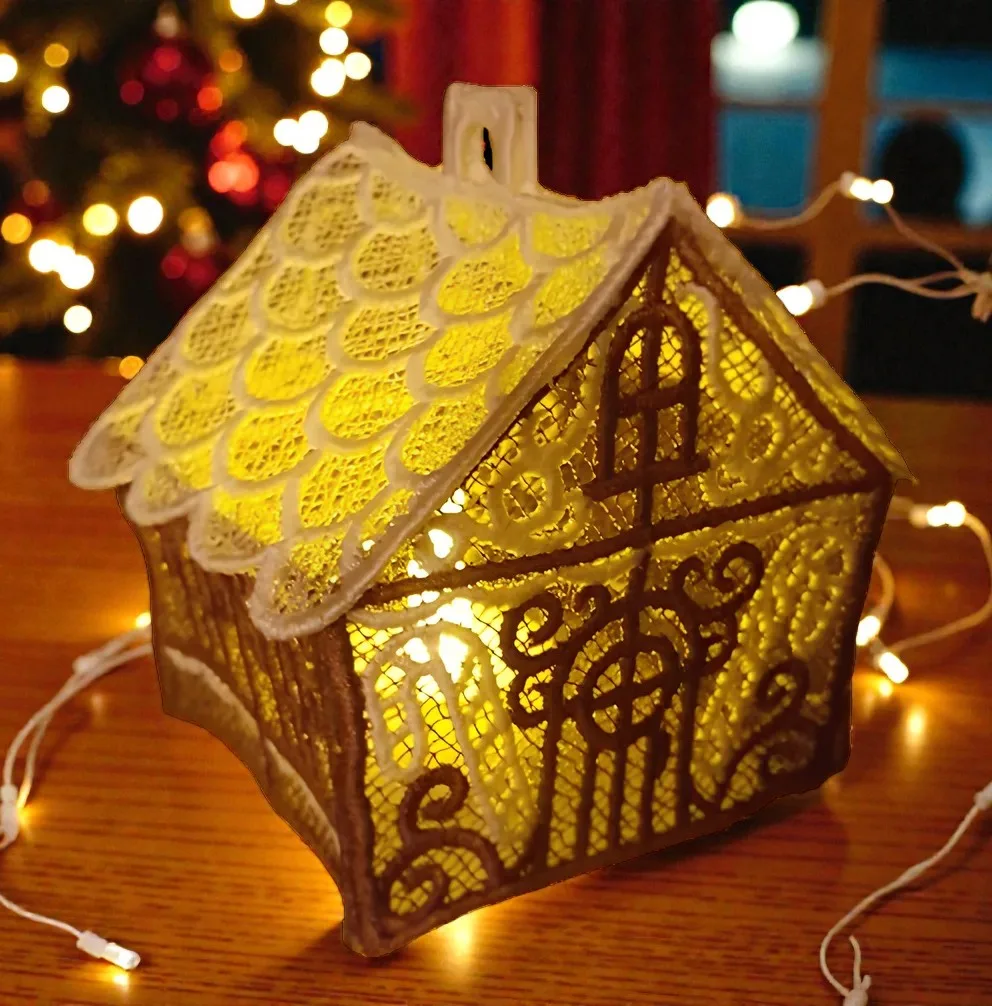 Gesticktes Lebkuchenhaus aus Spitze in Creme und Braun mit gelben Lichteffekten auf Holztisch.